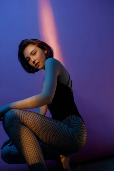 young woman sitting in black bodysuit and fishnet tights in squat pose on violet and pink background