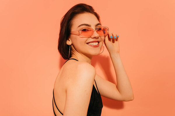 smiling young woman posing, looking at camera and adjusting sunglasses on orange background