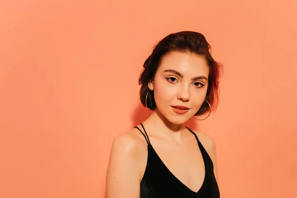 Siyah askılı kısa saçlı, turuncu arka planda genç bir kadın.
