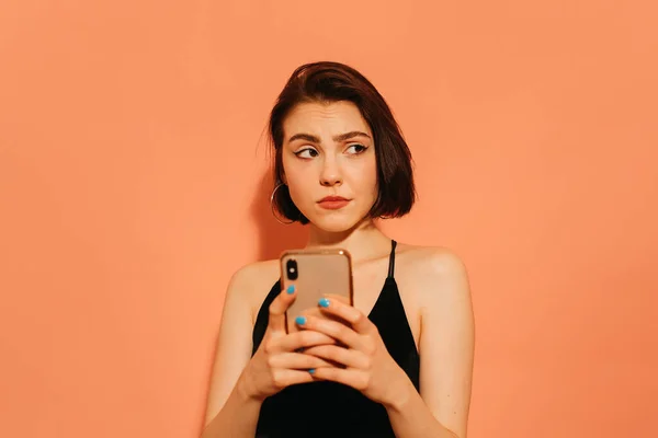 Elinde cep telefonu olan, turuncu arka planda siyah bir atlet giyen gergin genç bir kadın.