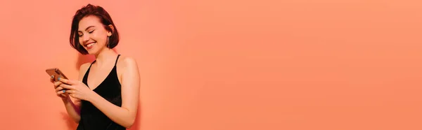 Genç bir kadın elinde akıllı telefona bakıyor ve turuncu arka planda gülümsüyor. 