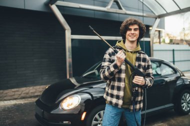 Gülümseyen sürücü oto yıkama makinesinin yanında oto yıkama makinesi tutuyor.
