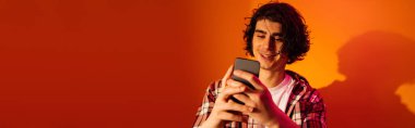 Turuncu arka planda cep telefonu kullanan neşeli kıvırcık adam, afiş 