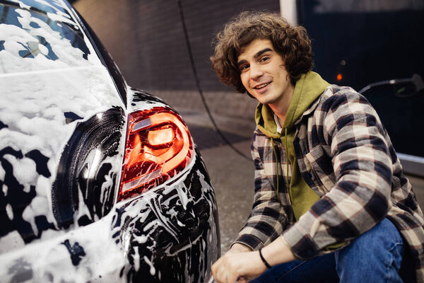 Young man smiling at camera near car in detergent on self service wash