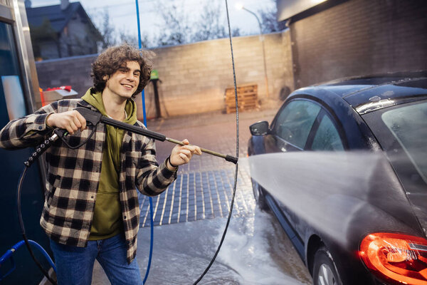 Cheerful man with pressure washer cleaning auto on self service wash in evening 