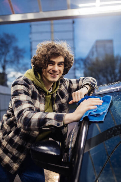 Curly man smiling at camera while cleaning wet car outdoors in evening 