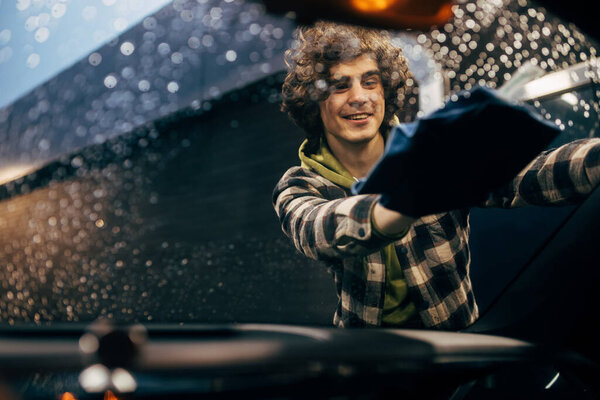 Smiling driver cleaning windshield of car with rag in evening 