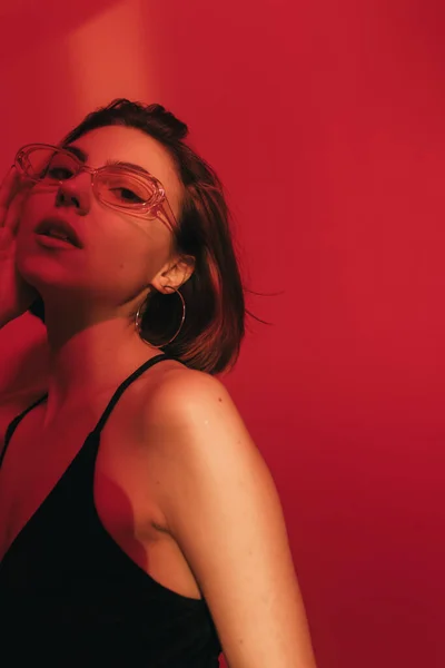 Young woman in black tank top and sunglasses looking at camera in dramatic light on red background — Stock Photo