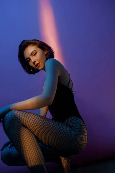 Young woman sitting in black bodysuit and fishnet tights in squat pose on violet and pink background — Stock Photo