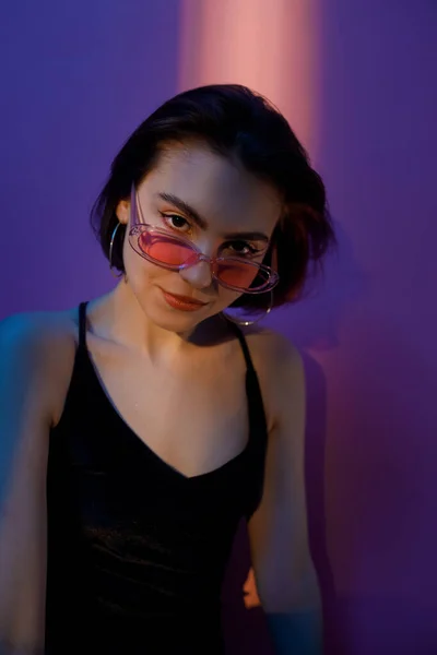 Young adult woman in black tank top and sunglasses looking at camera on violet and pink background — Stock Photo