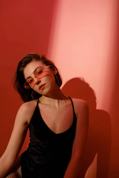 Sensual jovem mulher em preto bodysuit e óculos de sol olhando para a câmera no fundo vermelho com sombras — Fotografia de Stock