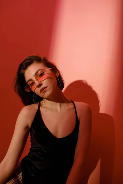 Sensual mujer joven en traje de cuerpo negro y gafas de sol mirando a la cámara sobre fondo rojo con sombras - foto de stock