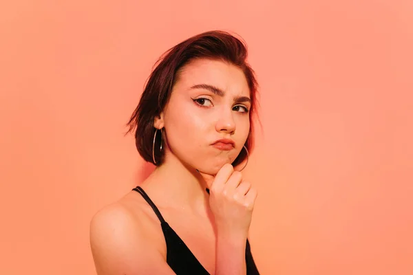 Mujer joven reflexiva mirando hacia otro lado con la mano cerca de la cara sobre fondo naranja - foto de stock