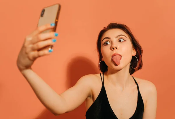 Junge Frau macht Selfie und grimassiert mit herausgestreckter Zunge auf orangefarbenem Hintergrund — Stockfoto