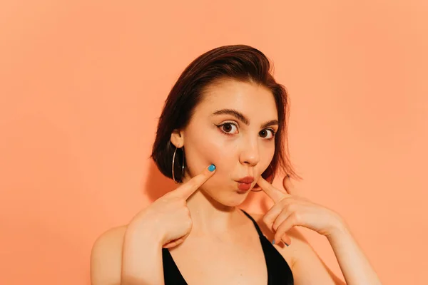 Young woman looking at camera and grimacing with pouting lips and hands near face on orange background — Stock Photo