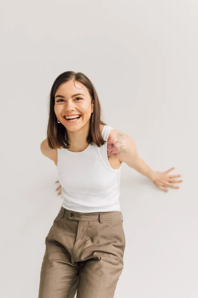 Mujer joven riendo en top blanco y pantalones beige sobre fondo gris - foto de stock