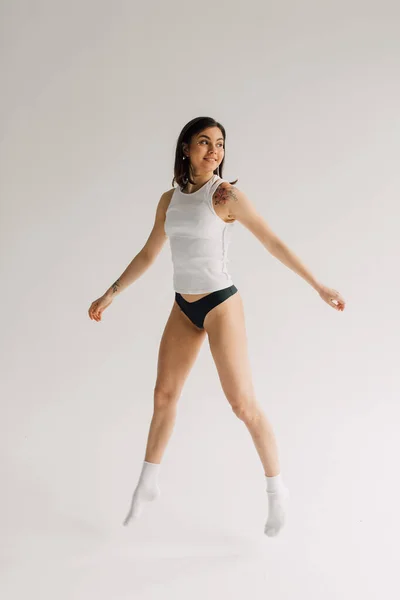 Fit young woman in white top, socks and black panties levitating on grey background — Stock Photo