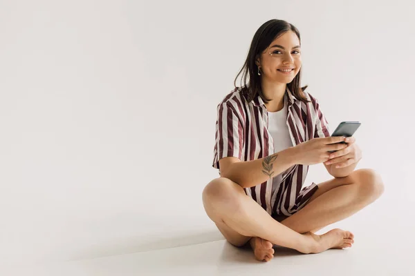 Положительная молодая женщина в белом топе и полосатой рубашке сидит со скрещенными ногами и держа смартфон на сером фоне — стоковое фото