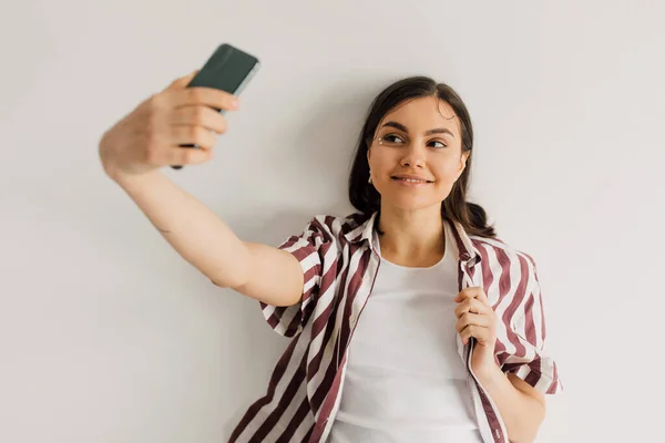 Положительная молодая женщина в полосатой рубашке делает селфи со смартфоном на сером фоне — стоковое фото