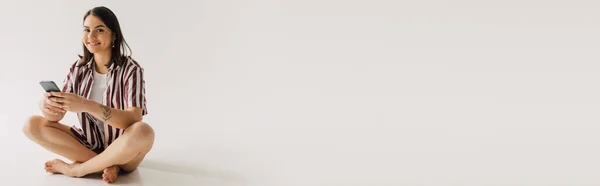 Позитивная молодая женщина в белом топе и в полосатой рубашке сидит в скрещенных ногах поза со смартфоном на сером фоне, баннер — стоковое фото