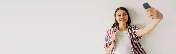Позитивная молодая женщина делает селфи со смартфоном на сером фоне, баннер — стоковое фото