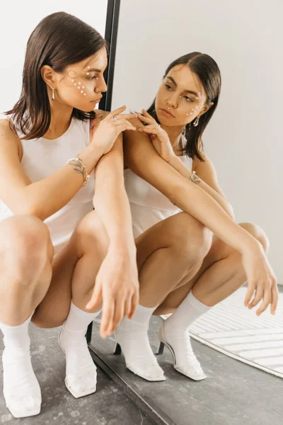 Молода стильна жінка з декоративним макіяжем, що сидить у присілій позі біля дзеркала з відображенням — стокове фото