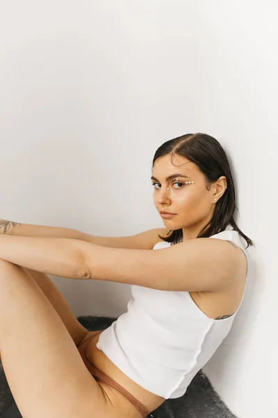 Young brunette woman sitting on the floor and looking at camera on grey background — Stock Photo