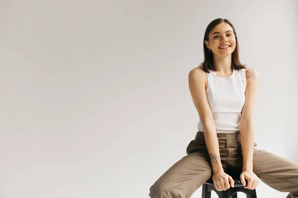 Young smiling woman in white top and beige trousers sitting on stool on grey background — Stock Photo