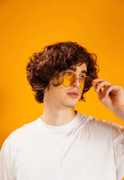 Young curly man in white t-shirt holding sunglasses isolated on orange — Stock Photo