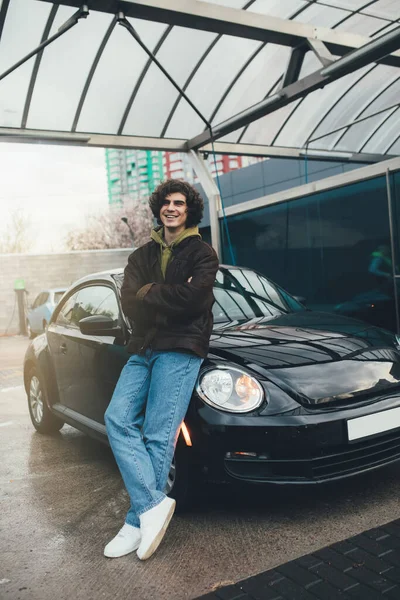 Homem alegre de pé com braços cruzados perto de carro preto na lavagem de auto-serviço — Fotografia de Stock