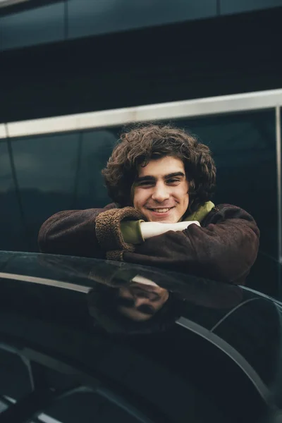 Hombre rizado sonriendo a la cámara cerca de la parte superior del coche negro al aire libre - foto de stock