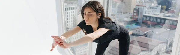 Young armenian woman in earphone doing forward lunges near window, banner — Stock Photo