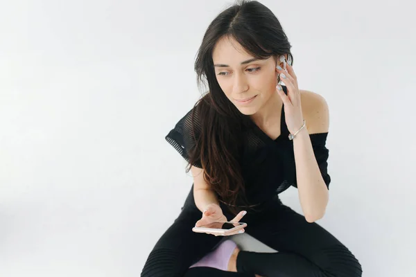 Armenian woman in black clothes adjusting earphone while sitting with mobile phone on white — Stock Photo