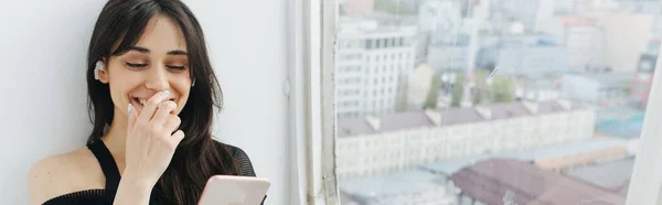 Feliz mujer armenia sonriendo mientras mira el teléfono inteligente cerca de la ventana, bandera - foto de stock