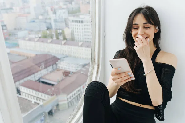 Heureuse femme arménienne couvrant la bouche avec la main tout en utilisant smartphone près de la fenêtre — Photo de stock
