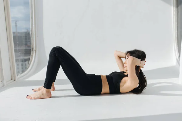 Jeune femme arménienne en vêtements de sport noirs pratiquant le yoga sur sol blanc près de la fenêtre — Photo de stock