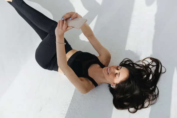 Overhead view of smiling armenian woman in black top and leggings using smartphone while lying on white floor — Stock Photo