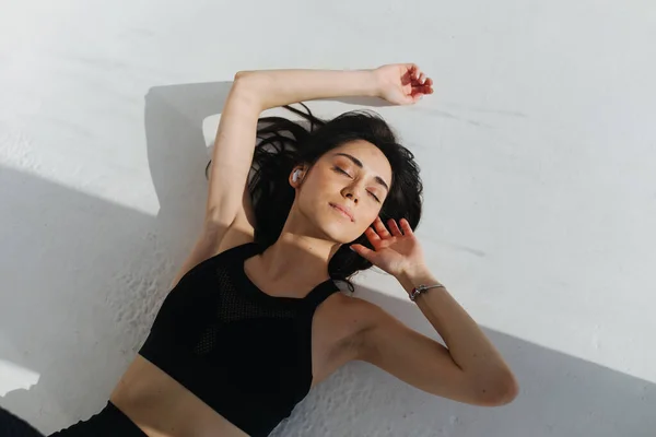 Top view of pretty armenian woman in wireless earphone relaxing on white floor with shadows — Stock Photo