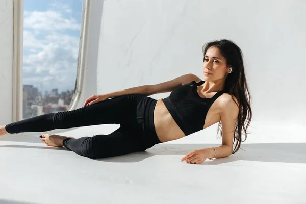 Стройная армянка в черной спортивной одежде медитирует на белом полу возле окна — стоковое фото