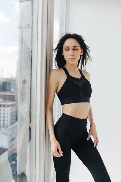 Slim armenian woman in black sportswear looking through window at home — Stock Photo