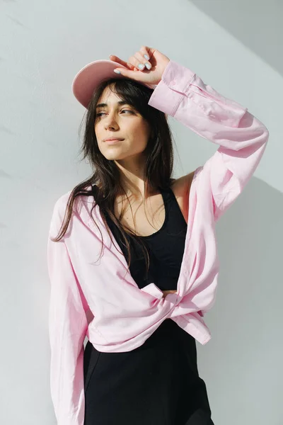 Bonita mujer armenia en camisa y gorra soñando cerca de la pared blanca con sombras - foto de stock