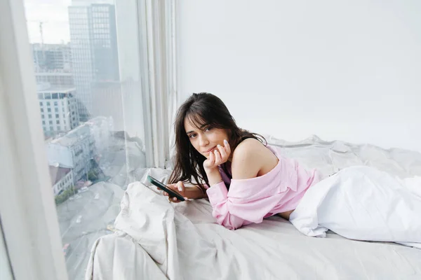 Bella donna armeno guardando la fotocamera mentre sdraiato su biancheria da letto bianca con smartphone — Foto stock