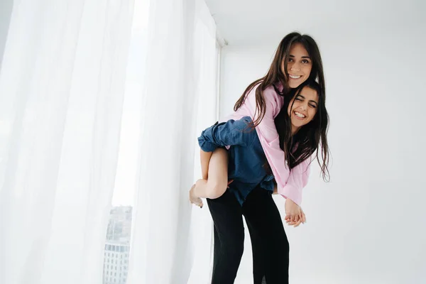 Excited armenian women piggybacking happy friend near window at home — Stock Photo