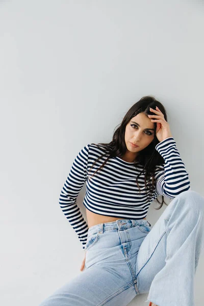 Brunette armenian woman in striped pullover and jeans sitting on white — Stock Photo