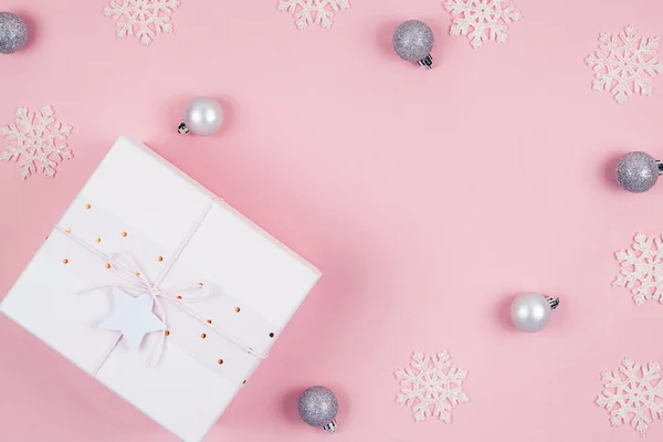 Gümüş süslemeli Noel hediyesinin üst görüntüsü toplar ve beyaz kar taneleri konfetisi pastel pembe arka planda..