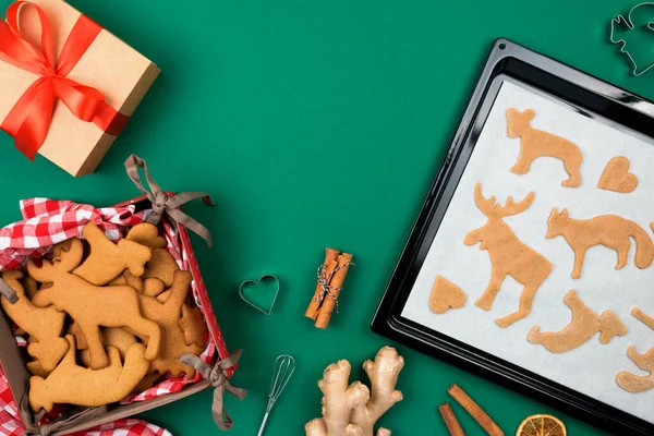 Vista superior de galletas de pan de jengibre de Navidad de masa cruda en bandeja para hornear, jengibre, canela, galleta con forma de ciervo, galletas recién respaldadas en servilleta roja a cuadros, regalo sobre verde. Puesta plana. — Foto de Stock