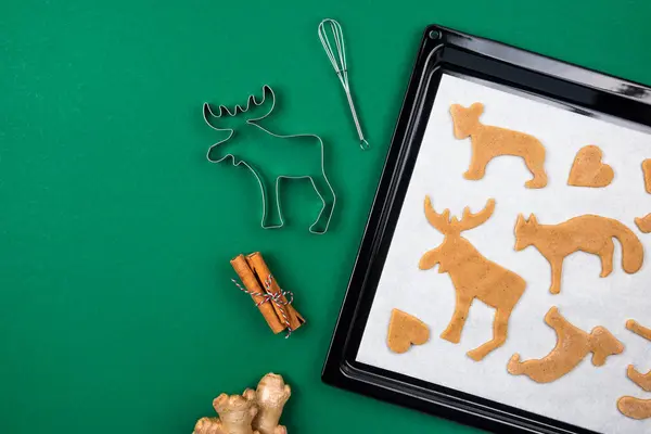 Vista superior de galletas de jengibre de Navidad de masa cruda sobre papel pergamino blanco bandeja para hornear en bandeja, jengibre, canela, cortador de galletas de ciervo para dar forma a la masa sobre verde — Foto de Stock