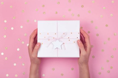 Altın konfetiyle süslenmiş pembe hediye kutusuyla serçe parmak arkaplanının üzerinde yıldız ve yay tutan kadın elleri. Düz konsept bayram şenliği.