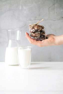 Ön tarafta taze pişmiş çikolatalı kurabiyeler ve bir bardak sütü ahşap masada tutarken görülüyor.