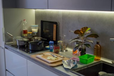 Tencereler içinde kepçeyle sobanın üzerinde duruyor, tahtanın içinde doğranmış soğan ve bıçak var, folyo yakınlarda yatıyor, tuzluk, bardak, rende, yağ şişesi, bitki, lavabo, askı ve sünger tam pişirme alanı.